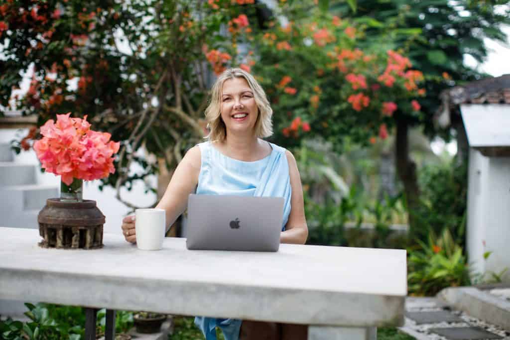 Kristina smiling outdoors in a garden, sitting at a table with her laptop and a cup of coffee, offering online life coaching