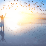 Woman standing on a mirror-like salt flat at sunrise with her arms raised, birds scattering into the light