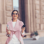 Woman in a pink coat walking through a city street, smiling, holding her phone
